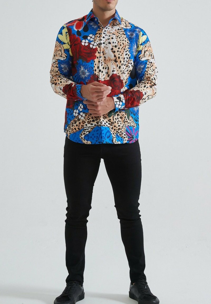 Chemise à manches longues colorée présentant un mélange d'imprimés animaliers et de motifs floraux, principalement dans des tons de bleu, rouge et crème, avec une fermeture à boutons.