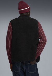 Schwarze Fleece-Weste mit weicher Textur, getragen über einem maroonfarbenen gestreiften Langarmshirt. Eine rote Strickmütze vervollständigt das Outfit.