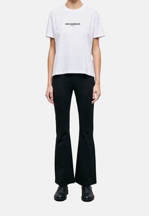 Persona con camiseta blanca de manga corta con el texto "The Kooples Paris", pantalones negros acampanados y zapatos negros sobre un fondo liso.