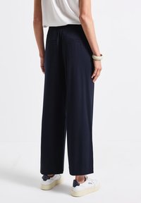 Marineblauwe wijde broek met elastische tailleband, twee achterzakken en een gladde stoftextuur, gecombineerd met witte sneakers en een beige armband.