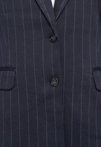 Veste de costume bleu marine foncé à fines rayures avec deux boutons noirs et deux poches à rabat visibles à l'avant.