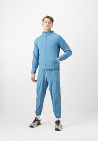 Lyseblå Nike træningsdragt med en lynlåsjakke med lommer og elastiske manchetter, parret med matchende bukser med elastiske manchetter og sportssko.