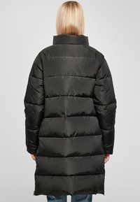 Giacca a vento nera con silhouette lunga, caratterizzata da sezioni trapuntate orizzontali, un colletto alto e una superficie liscia e lucida.