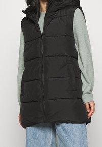 Svart puffväst med huva, med horisontell vaddering, en dragkedja framtill och sidofickor, bärs över en ljusgrön tröja.