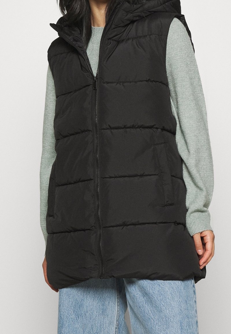 Svart puffväst med huva, med horisontell vaddering, en dragkedja framtill och sidofickor, bärs över en ljusgrön tröja.