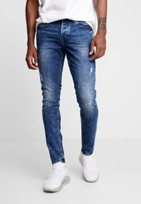 Slim-fit blue denim jeans with distressed details and a faded finish. Features standard pockets and belt loops, paired with white sneakers.