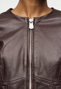 Κοντινό πλάνο ενός ατόμου που φοράει σκούρο καφέ δερμάτινο μπουφάν με ασημένιο μπροστινό φερμουάρ, το οποίο είναι μερικώς κλειστό κοντά στην περιοχή της κλείδας.