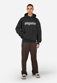 Μαύρη φούτερ μπλούζα με κείμενο "pegador" σε γκρι, διαθέτει τσέπη καγκουρό και ριμπ μανσέτες. Συνδυάζεται με καφέ παντελόνι και αθλητικά παπούτσια.