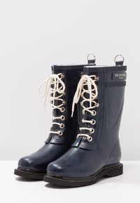 Bottes de pluie en caoutchouc bleu marine avec des lacets blancs et des œillets en argent. Bout arrondi, semelle épaisse noire et accents en tissu en haut avec un détail de logo.