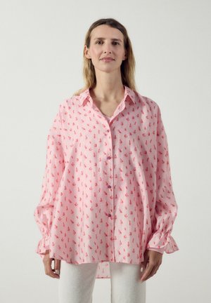 Frau trägt ein locker sitzendes, rosa Hemd mit roten Segelboot-Mustern und weiße Hose, steht vor einem schlichten hellen Hintergrund.