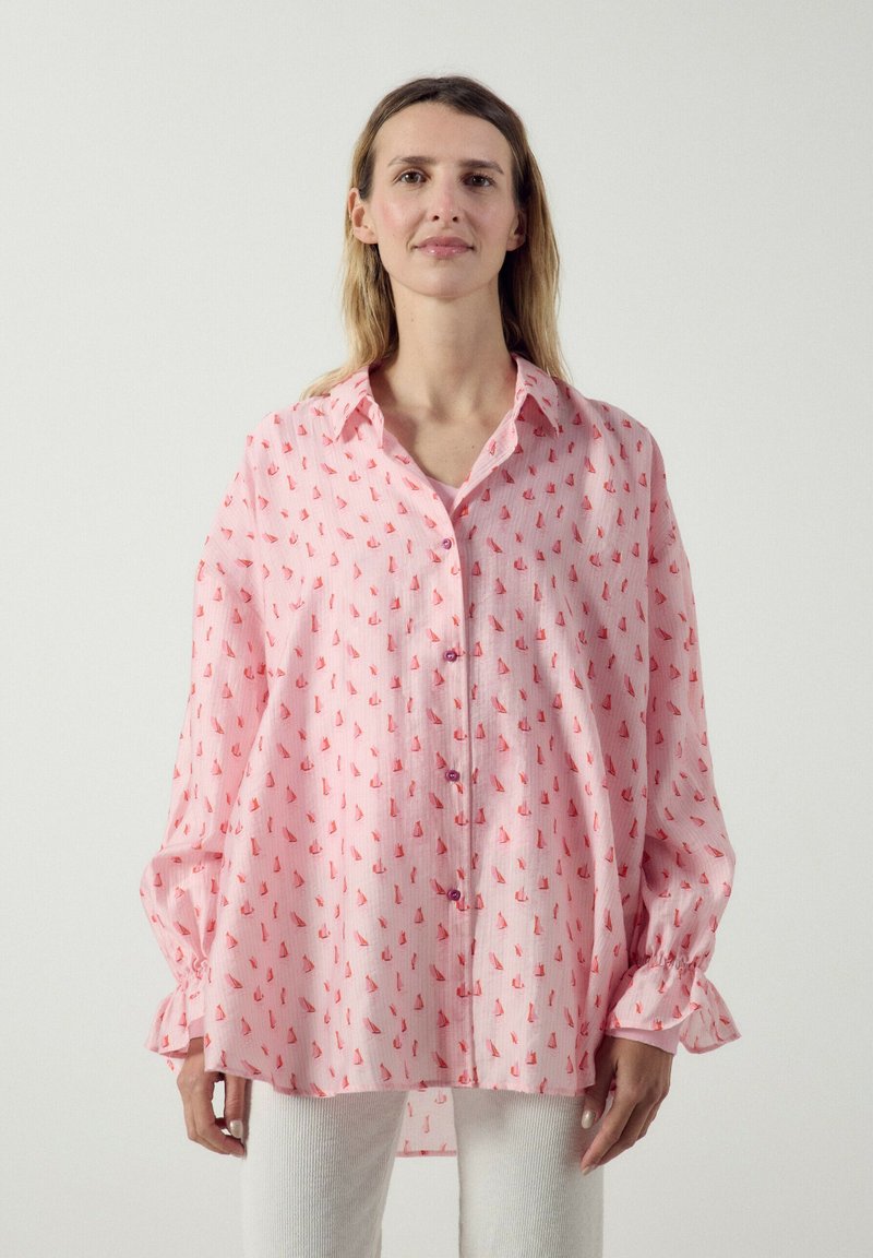 Femme portant une chemise rose ample à boutons avec des motifs de voiliers rouges et un pantalon blanc, debout devant un fond clair uni.