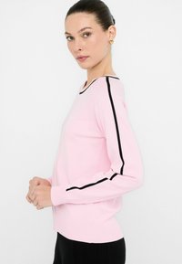 Frau trägt einen hellrosa Pullover mit schwarzen Streifen an den Ärmeln und am Ausschnitt, kombiniert mit schwarzen Hosen, vor einem schlichten weißen Hintergrund.