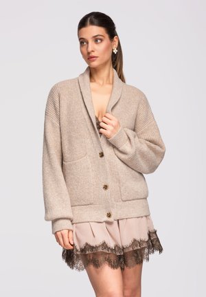 Mujer con cabello castaño recogido en una coleta lleva un cárdigan de punto beige sobre un vestido rosa claro con detalles de encaje negro, mirando ligeramente hacia un lado.