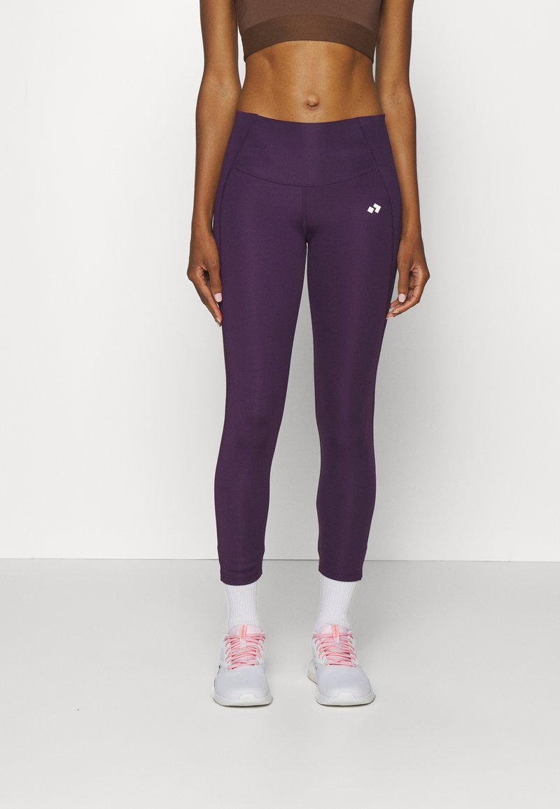 Fialové sportovní legíny s hladkou texturou, vysokým pasem a malým logem na stehně. V kombinaci s bílými ponožkami a světlými teniskami.