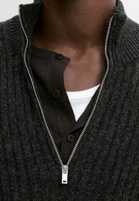 Gros plan sur une personne portant un pull zippé gris foncé par-dessus une chemise foncée boutonnée et un sous-vêtement blanc.