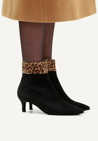 Des bottines en suédine noire avec un bout pointu et un revers imprimé léopard ; le talon est bas et effilé, offrant une silhouette élégante.