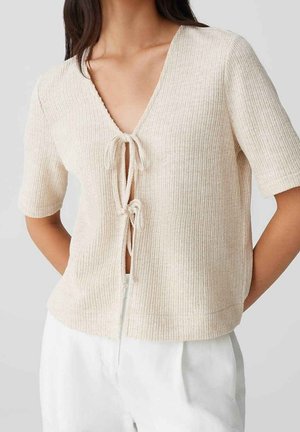 Haut en maille côtelée beige clair à manches courtes avec deux fermetures par nœuds à l'avant, porté avec un pantalon blanc, sur une personne debout devant un fond uni.