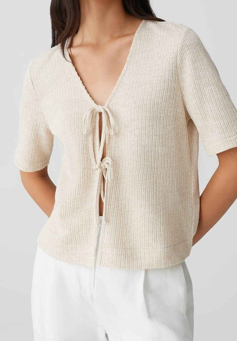Haut en maille côtelée beige clair à manches courtes avec deux fermetures par nœuds à l'avant, porté avec un pantalon blanc, sur une personne debout devant un fond uni.