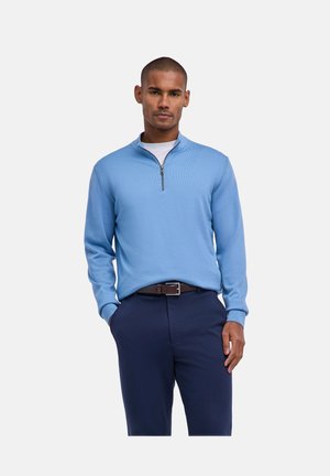 Homme portant un pull bleu clair à fermeture éclair à quart de zip sur une chemise blanche, pantalon bleu marine et ceinture marron, debout avec une main dans la poche.