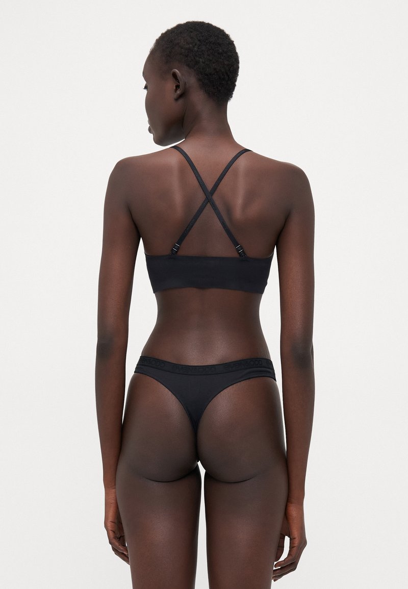 Bralette sans fil noir avec des bretelles croisées et un tissu doux. String noir assorti avec une bande logo à la taille. Texture lisse.