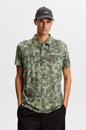 Mann trägt ein grün-braunes, pixeliertes Tarn-Poloshirt und eine schwarze Kappe, Hände in den Taschen, neutraler Hintergrund.