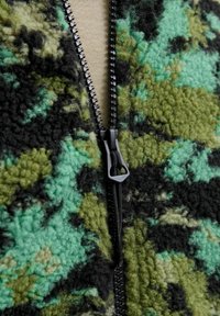 Fleece jack in een camouflagemuster van donkergroen, lichtgroen en zwart met een zwarte rits. De textuur is zacht en fuzzy, met een robuuste uitstraling.