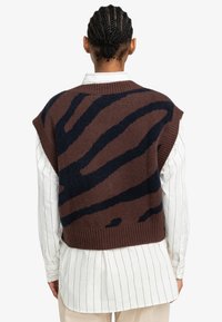 Ärmelloser gestrickter Pullover mit einem braun-blauen Zebra-Muster, geripptem Saum, oversized Schnitt, über einem weiß gestreiften Hemd getragen.