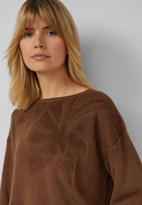 Pull tricot marron présentant un motif floral texturé, un large col et une coupe décontractée, avec une finition douce et des détails minimaux.