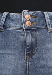 Denim jeans med middels høyt liv, to kopperknapper og kontrasterende oransje søm. Stoffet er lyseblått med en myk tekstur.