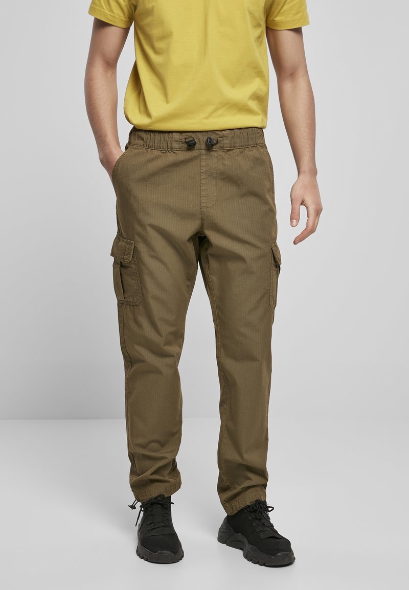 Homme portant un pantalon cargo marron avec des bas élastiques, associé à un t-shirt jaune et des baskets noires, debout avec une main dans la poche.