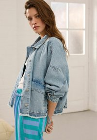 Veste en denim bleu clair avec une coupe décontractée, des épaules tombantes et un ourlet effiloché, assortie à un pantalon rayé teal et bleu.
