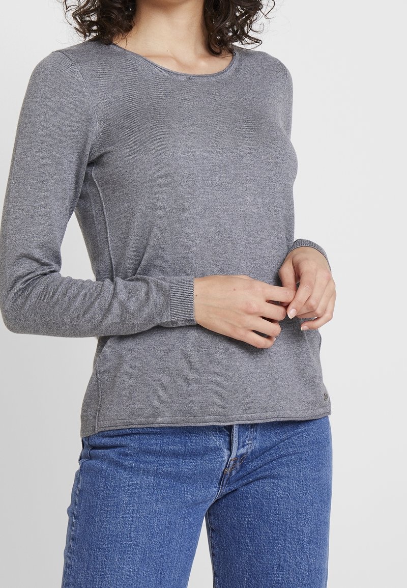 Grå langærmet top lavet af ribbet stof med rund halsudskæring. Har en tætsiddende design og subtile sømdetaljer. Bæres med blå jeans.