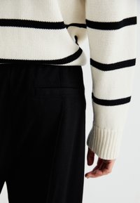 Personne portant un pull blanc à rayures horizontales noires et un pantalon noir, présentée de la taille à la main sur un fond uni.