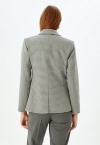Harmaa houndstooth-takkia lapelsolvilla, muotoiltu istuvuus, takana venttiilidetalji, yhdistetty harmaisiin housuihin, joissa on teksturoitu kangas.