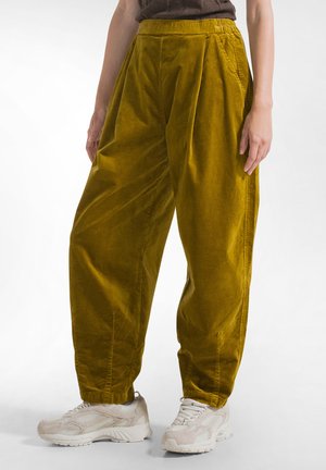 Persoon die losse mosterdgele corduroy broeken en witte stevige sneakers draagt, staand tegen een eenvoudige lichte achtergrond.