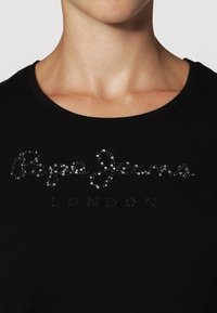 Nainen, joka on pukeutunut mustaan paita, jossa on pyöreä pääntie ja "Pepe Jeans London" -logo pienillä kiiltävillä niiteillä rinnassa.