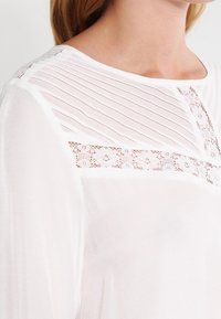 Blusa bianca con un plastron testurizzato, dettagli in pizzo e pieghe verticali sulle spalle. Tessuto leggero con scollatura rotonda.