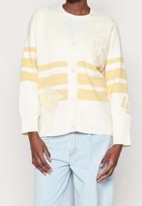Cardigan crema con righe gialle, scollo a V, quattro bottoni, toppa decorativa, texture morbida, polsini e orlo a coste. Indossato sopra jeans chiari.