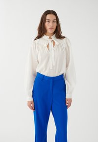 Blusa blanca con detalle de hombros fruncidos y cierre de lazo, combinada con pantalones de tiro alto de un brillante color azul, que presentan una textura suave.
