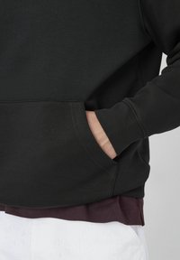 Sweatshirt noir avec poche kangourou, poignets côtelés et un ourlet bordeaux visible en bas. Texture douce et design simple.