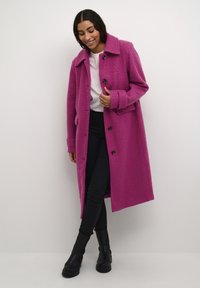Cappotto in peluche fucsia con colletto, bottoni frontali e tasche laterali. Indossato sopra una camicia bianca e abbinato a pantaloni neri e stivaletti alla caviglia.