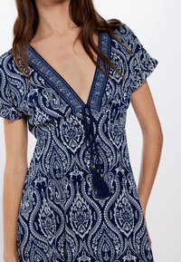 Robe bleu marine avec un motif paisley, col en V profond, manches courtes volantées, et un détail de franges nouées à l'avant. Tissu en coton, coupe décontractée.
