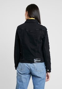 Chaqueta de mezclilla negra con un diseño ajustado, que presenta un parche con logo en la parte posterior, cierres de botón y costuras en contraste. Cuello plano y mangas largas.