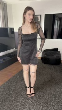 Melns piegulošs kombinezons ar caurspīdīgām garām piedurknēm un mirdzošu tekstūru. Kombinēts ar melnām augstpapēžu sandales ar spīļveida siksnām. Fonā redzams tumšs televizora plaukts.