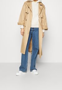 Cappotto trench beige realizzato in tessuto leggero, con un design doppiopetto, ampi revers e bottoni neri sopra un leggero felpa e jeans blu.