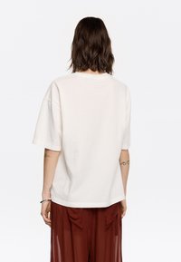 Übergroßes beiges Baumwoll-T-Shirt mit kurzen Ärmeln und Rundhalsausschnitt, präsentiert an einer Person, die leichte rotbraune Hosen trägt.