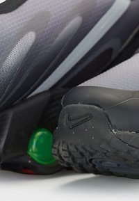 Buty sportowe z szarą cholewką z siateczki, czarną gumową podeszwą zewnętrzną, zieloną amortyzacją i charakterystycznym logo Nike na pięcie. Teksturowany, nowoczesny design.