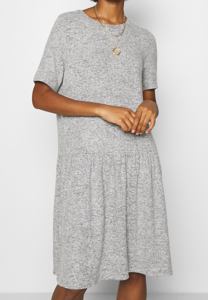 Femme portant une robe en maille grise à manches courtes avec taille basse et deux colliers dorés superposés, debout devant un fond blanc uni.