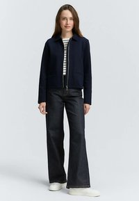 Laivastonsininen lyhyt vetoketjutakki, jossa on kaksi etutaskua ja kaulus, puettuna raidallisen paidan ja leveälahkeisten tummien farkkujen päälle, valkoisten tennareiden kanssa.