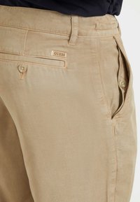 Pantalones de algodón beige con una textura suave, que cuentan con un bolsillo frontal, cierre de botón y un pequeño logo "GUESS" en la parte trasera.